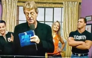 A screen capture from the Jerry Springer Show showing Jerry in the foreground, a man and a woman behind him in the background, and a burly security guy with a serious look on his face standing with shoulders crossed off to the side.